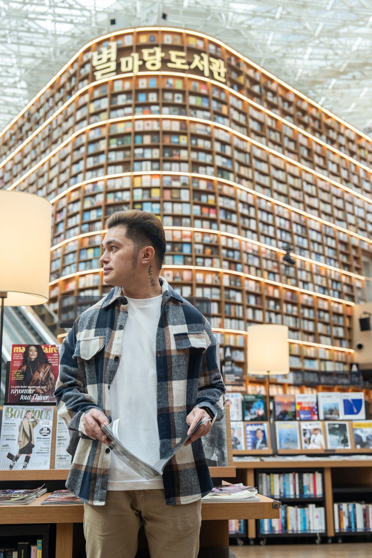 Man in plaid shirt stands in Starfield Library Seoul, surrounded by towering bookshelves filled with books.
