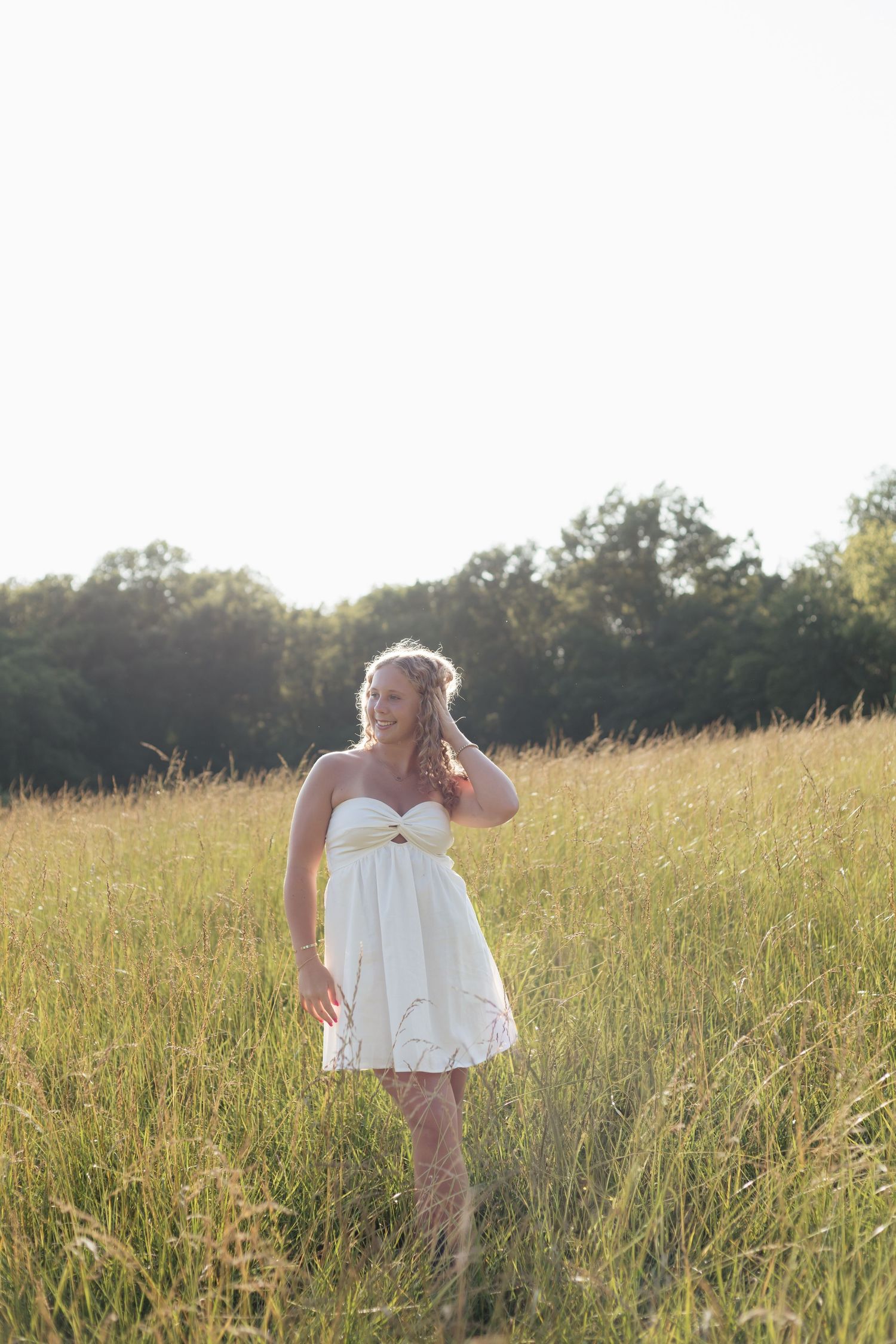 Young woman in white strapless dress and cowboy boots walking through golden meadow at sunset.