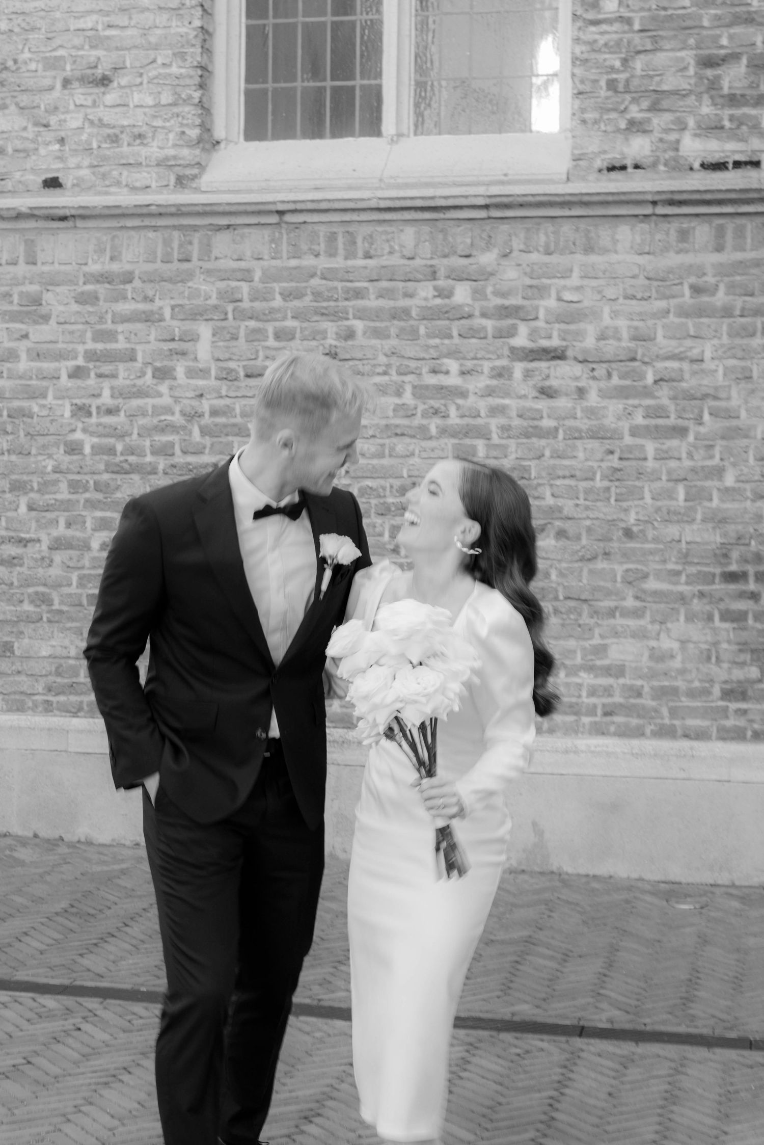 Black and white photo of a smiling bride holding white flowers and groom in tuxedo standing together near brick wall.