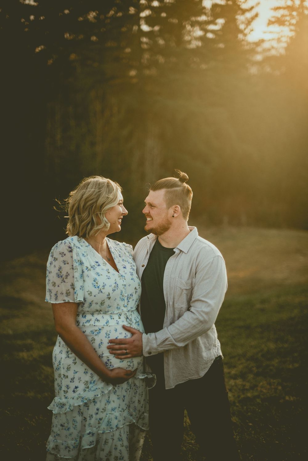 Couple sharing a tender moment outdoors at golden hour, man gently holding pregnant woman's belly in a sunlit forest setting.