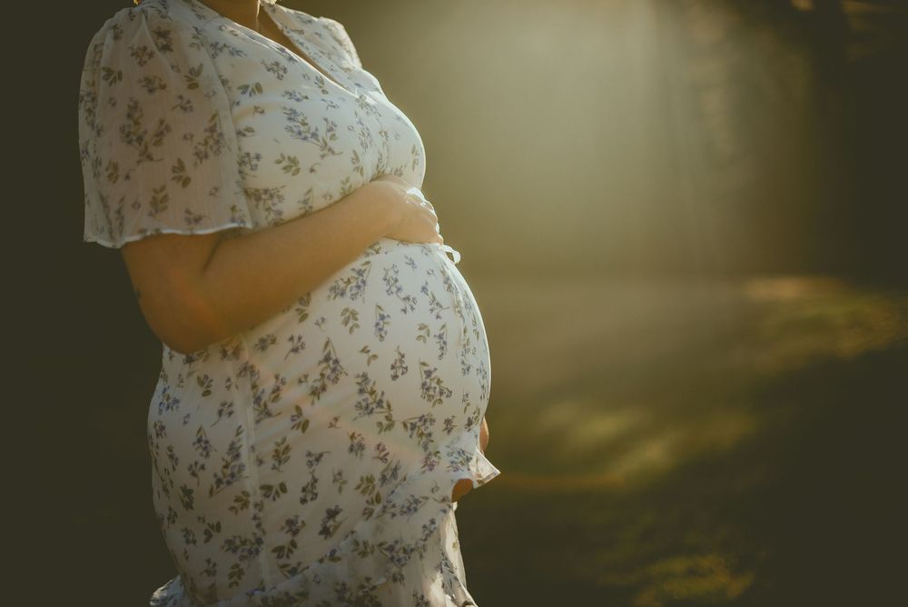 Pregnant woman in floral dress cradling baby bump outdoors in soft golden sunlight.