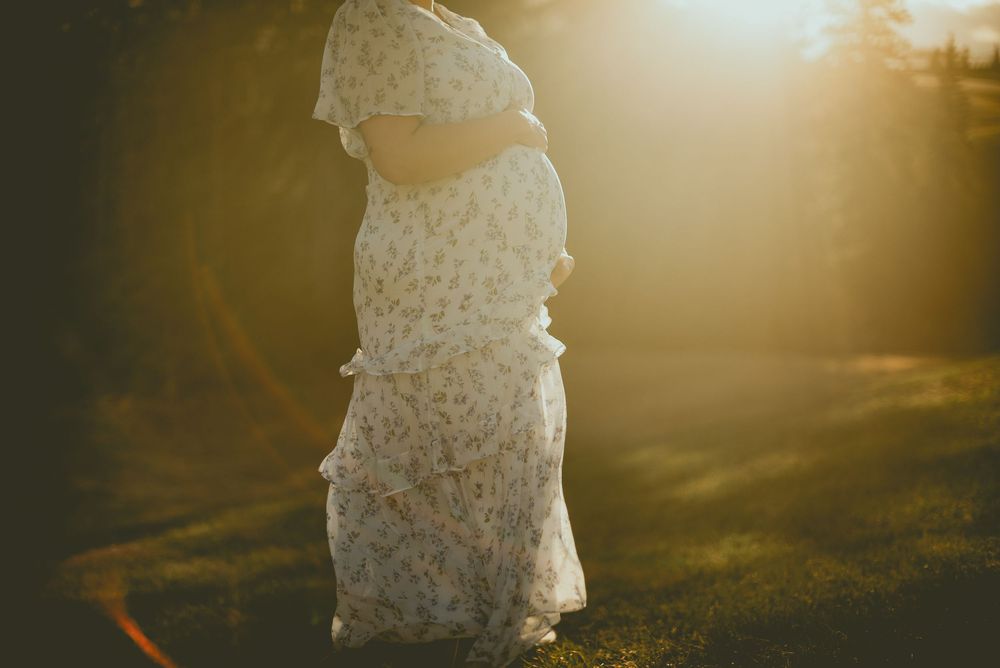 Pregnant woman in floral dress bathed in golden sunset light outdoors.