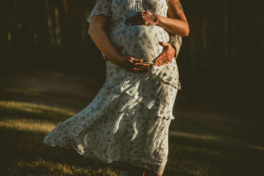 Pregnant woman in floral dress cradling baby bump outdoors in warm golden sunlight.