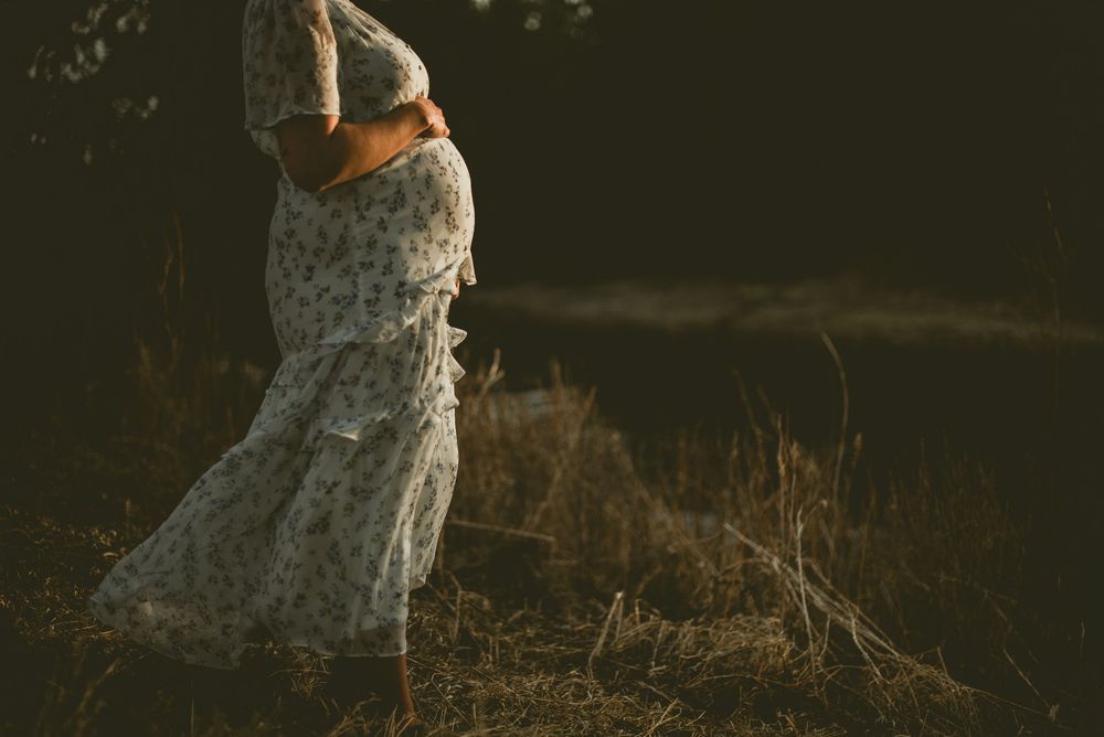 Pregnant woman in floral dress cradling baby bump outdoors in golden sunlight among dry grass.