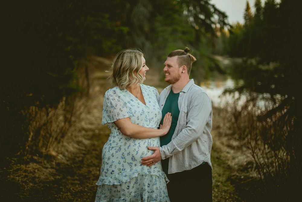 Couple smiling at each other outdoors, woman in floral dress cradling baby bump, man in green shirt and gray cardigan.