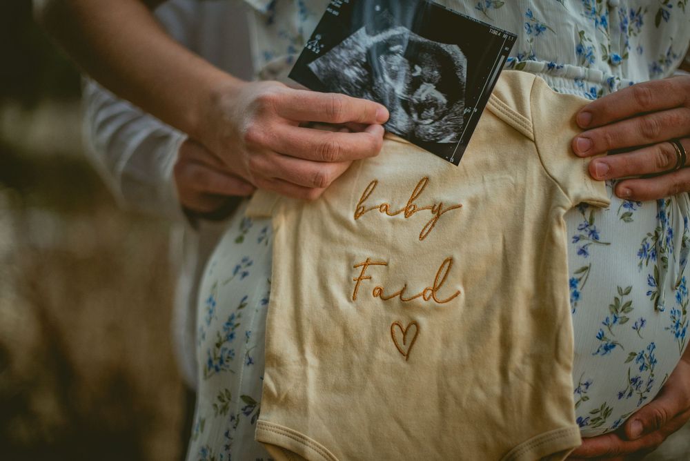 Couple holding ultrasound photo and baby onesie with name Ford during maternity announcement outdoors.
