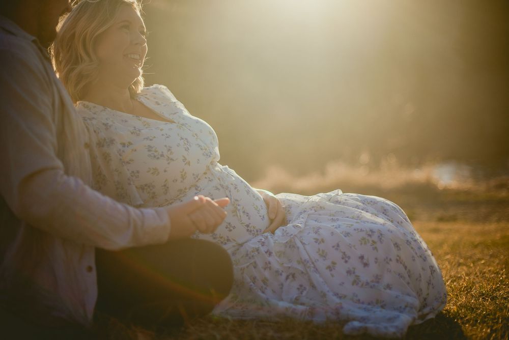 Pregnant woman in floral dress sitting with partner in golden sunlight during maternity photo session.