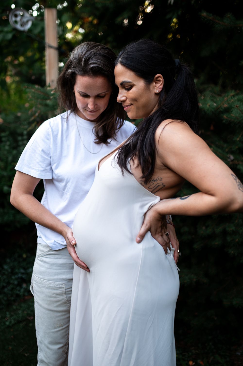 Two women in white outfits share a tender maternity moment outdoors, one gently cradling the pregnant belly.