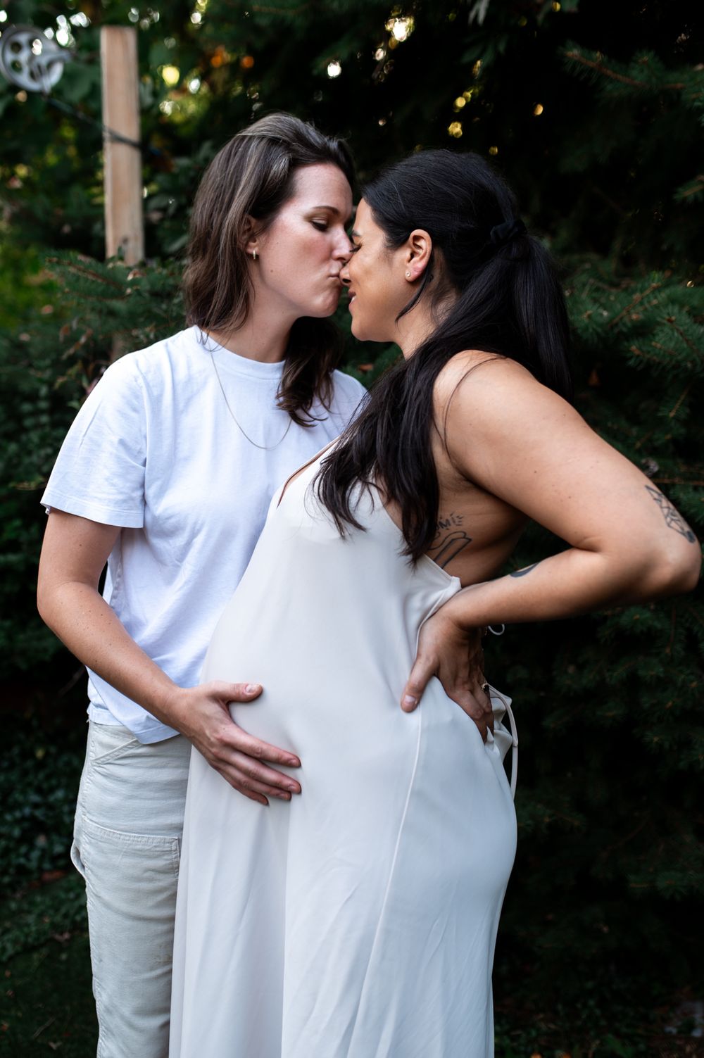Two women in white outfits sharing a kiss while tenderly cradling a baby bump outdoors.