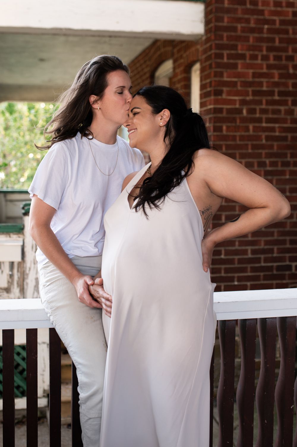 Two women in white outfits share a tender moment on a porch, one visibly pregnant.