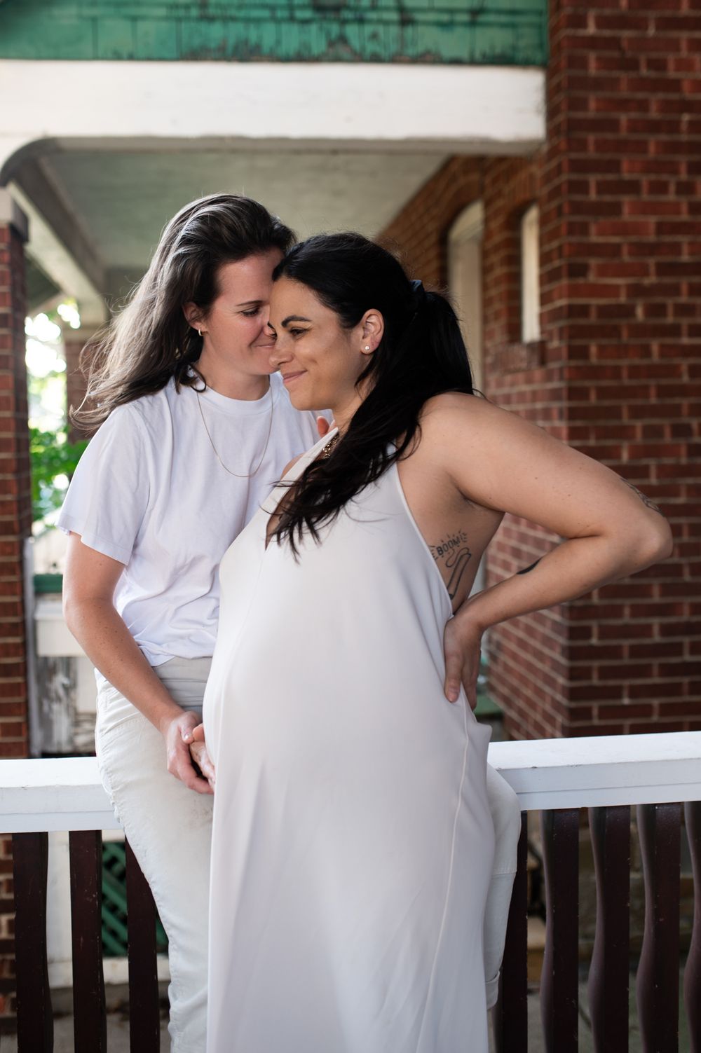 Two women in white outfits share a tender moment on a brick porch, one visibly pregnant.