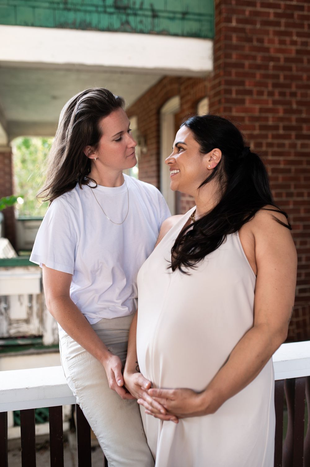 Pregnant woman in white dress smiles at partner on a sunlit porch during maternity photoshoot.