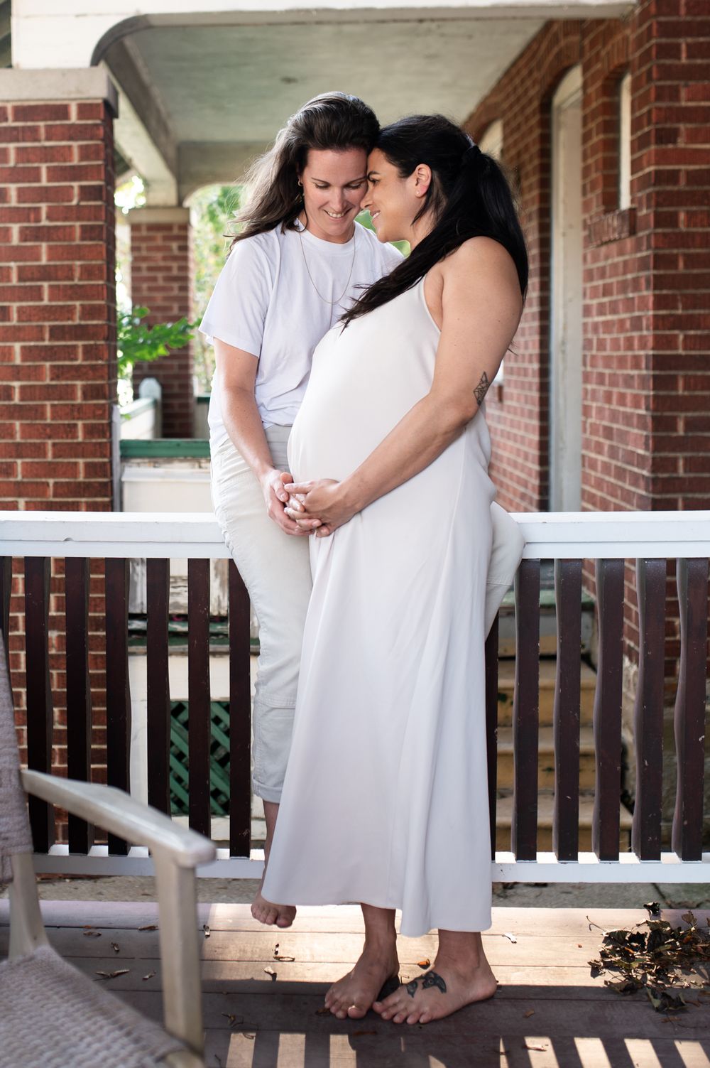 Two women embrace lovingly on a porch, one in a white maternity dress, sharing a joyful moment.