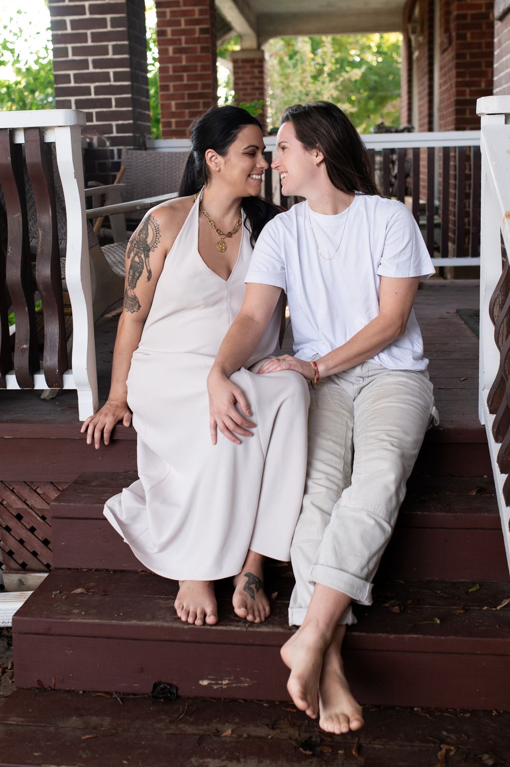 Two barefoot women in white sit on porch steps, smiling at each other during maternity session.
