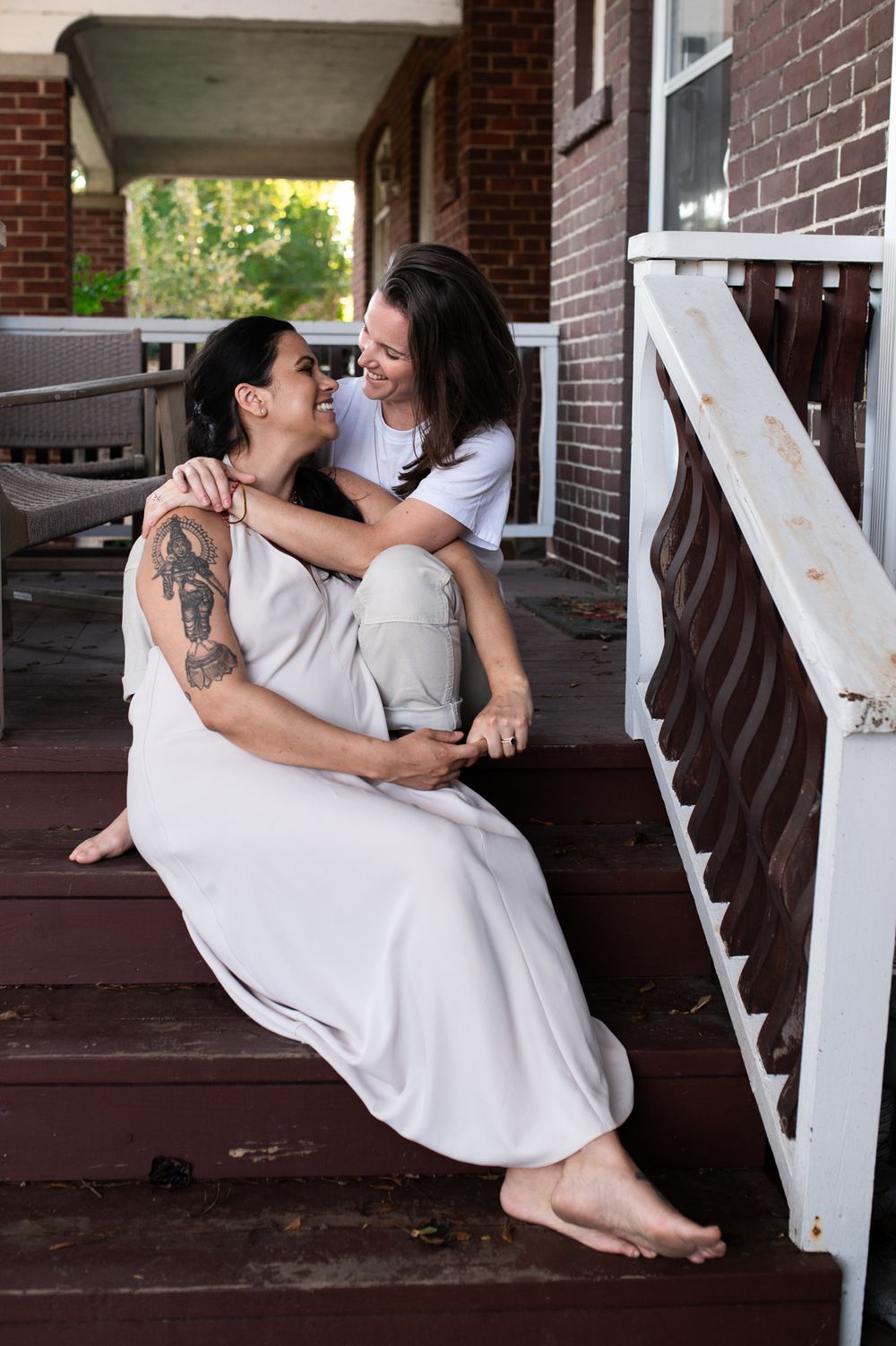 Two women share a joyful embrace while sitting on wooden porch steps in white outfits.