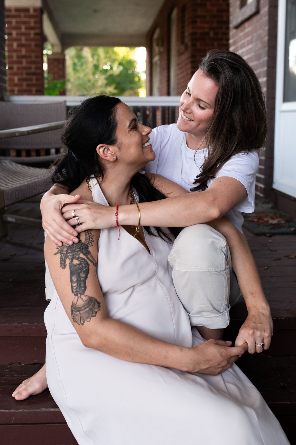Two women sharing a joyful embrace on a porch, one with a tattoo sleeve sitting on the others lap.