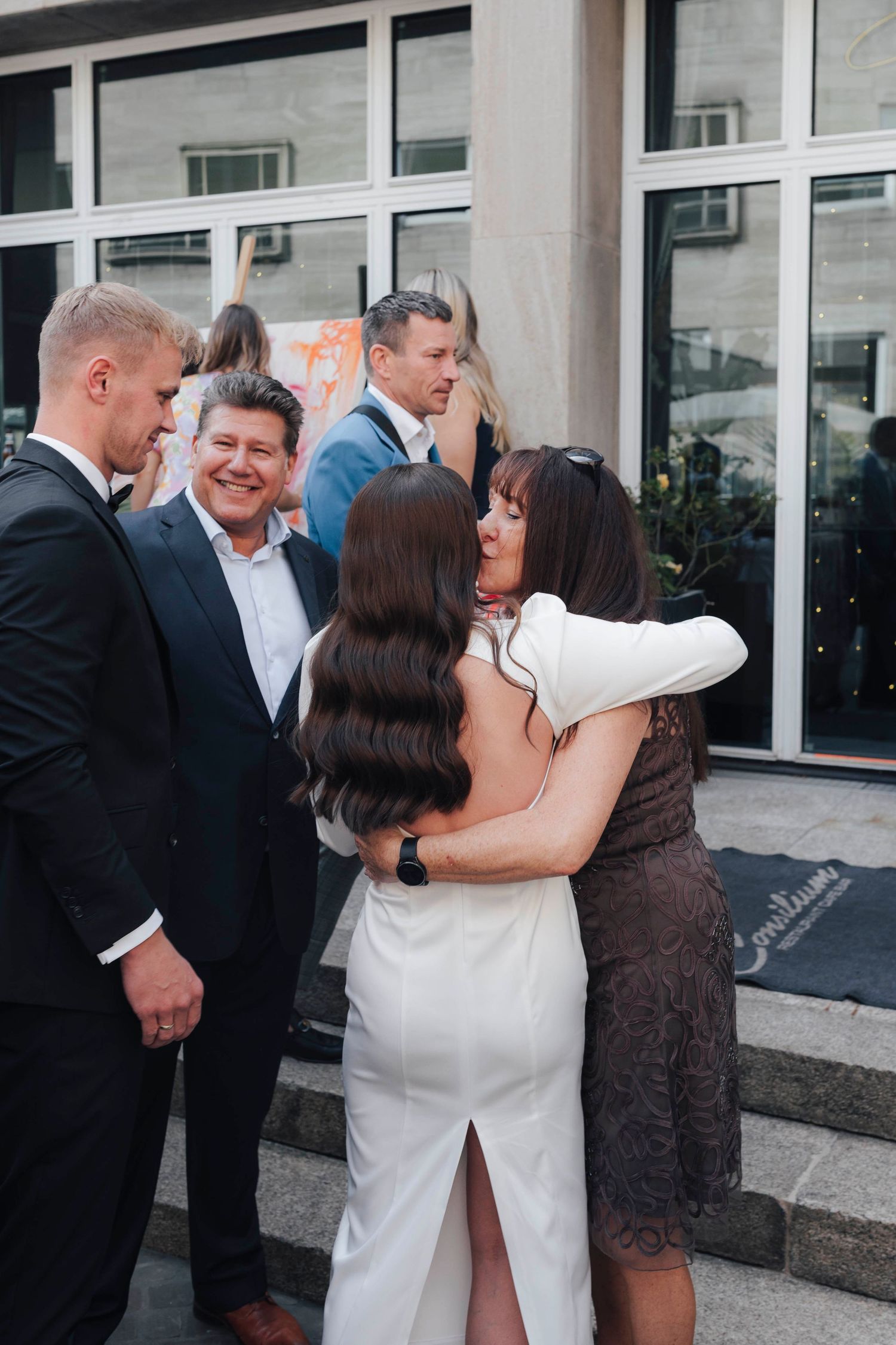 Bride in white dress hugging a guest at a wedding reception outside a building.