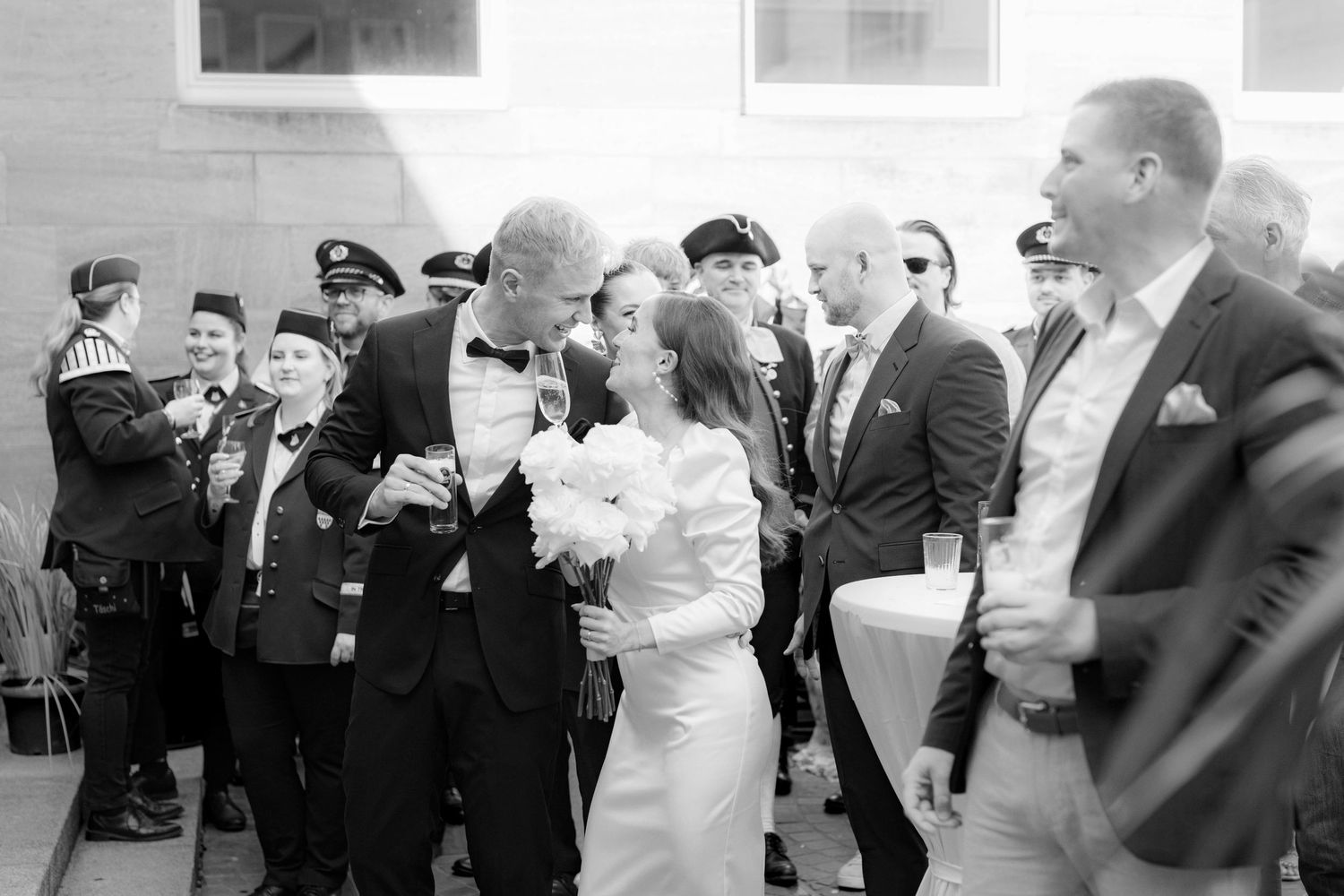 Black and white wedding photo of couple kissing surrounded by guests in military uniforms outdoors.