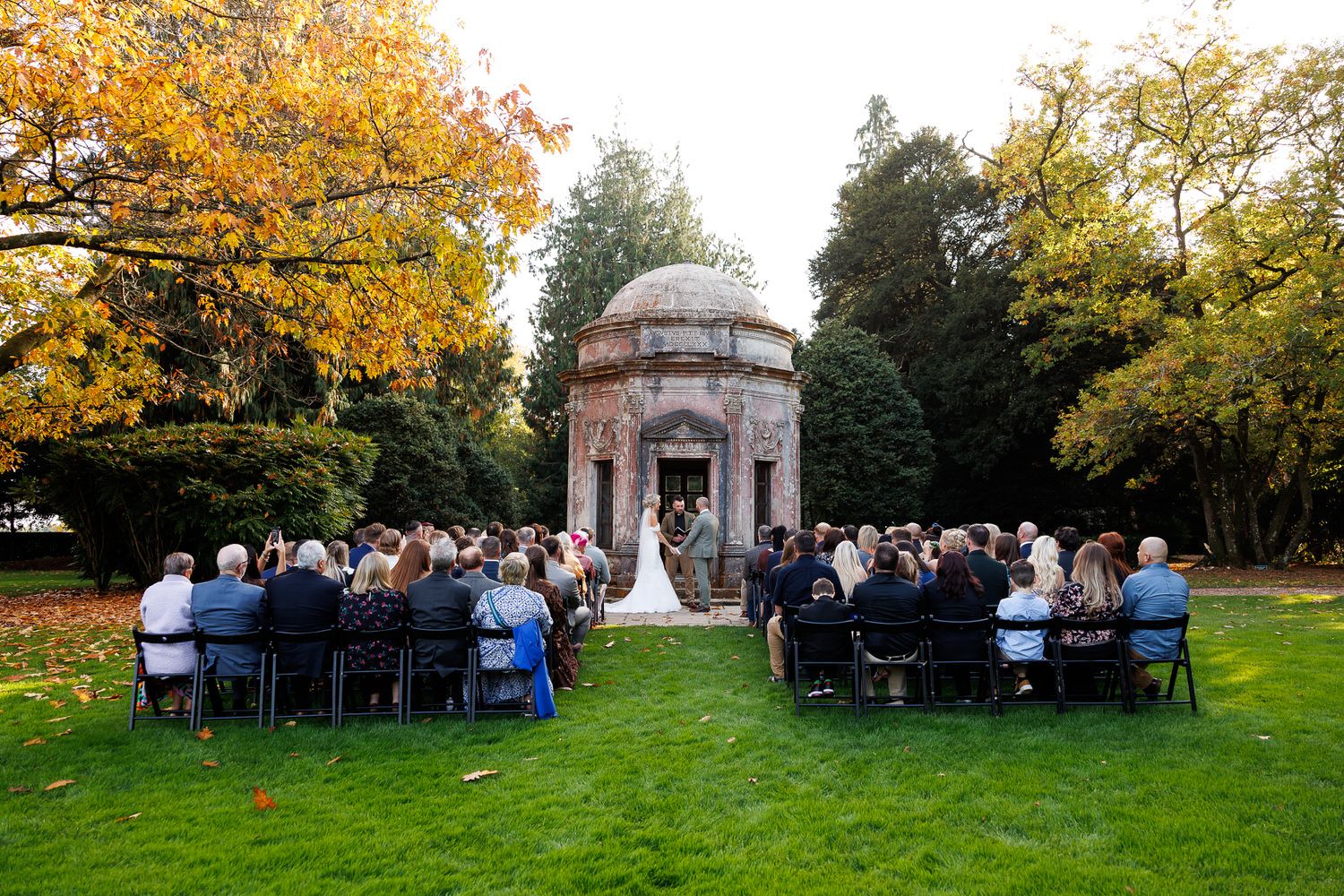 Outdoor wedding ceremony on green lawn before a historic stone domed mausoleum surrounded by autumn foliage.