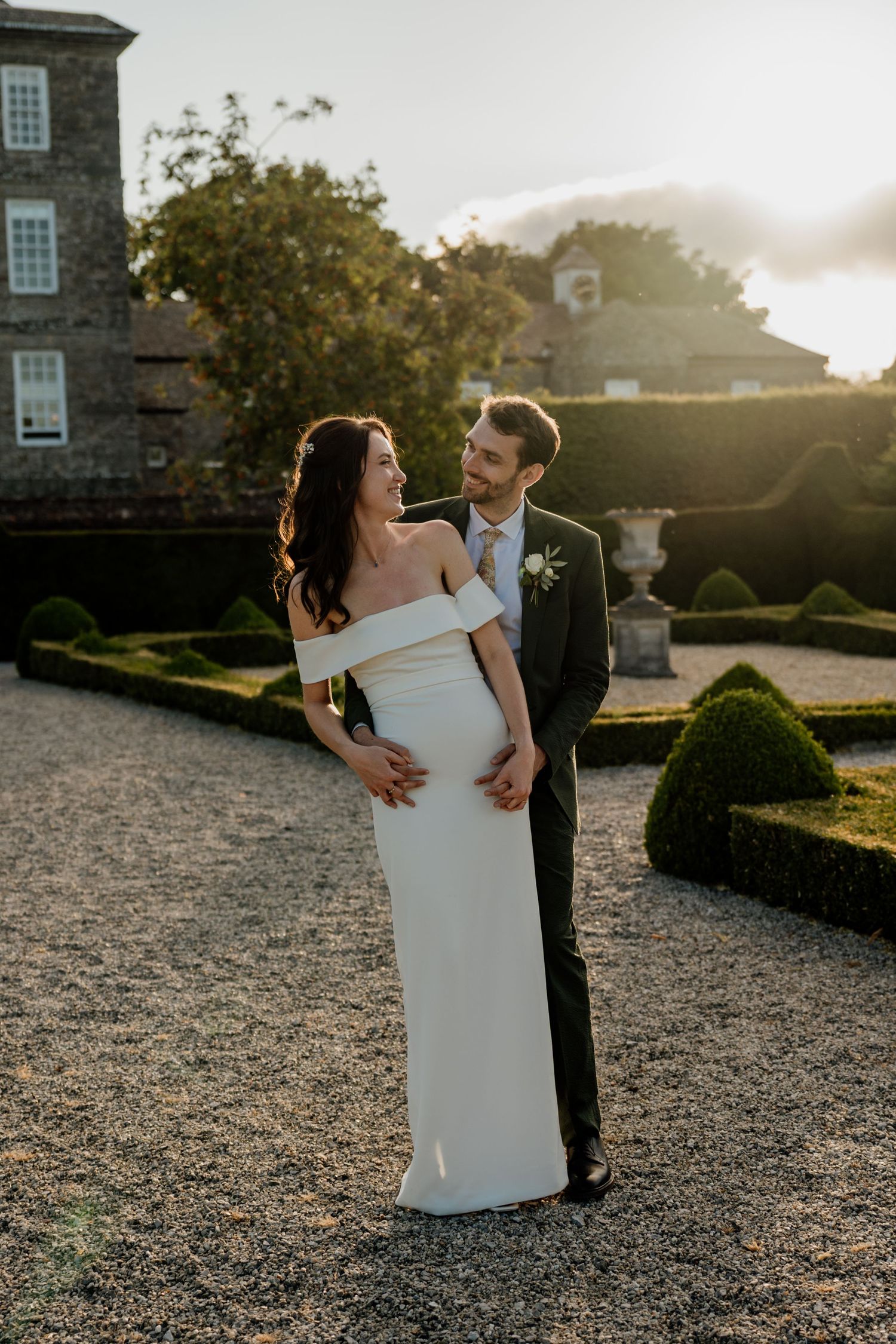A bride and groom share a romantic moment in a formal garden at golden hour, she wearing an off-shoulder white gown.