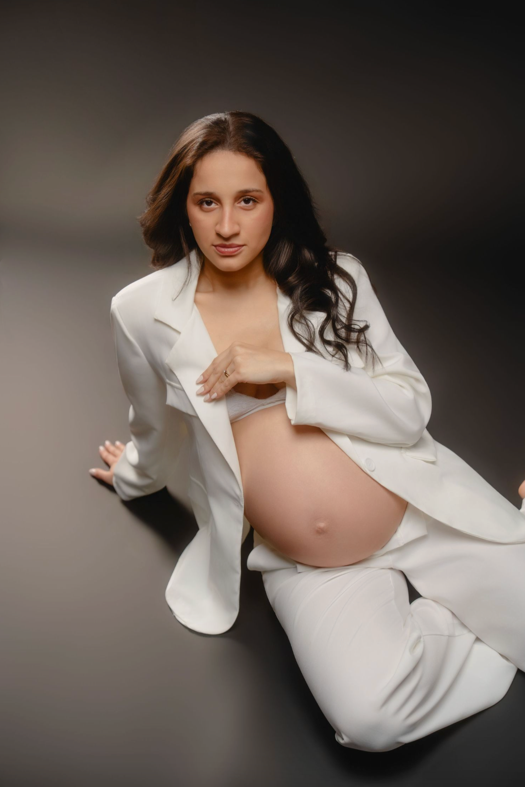 Pregnant woman in white blazer and pants sitting on floor, showcasing baby bump in elegant maternity photo shoot.