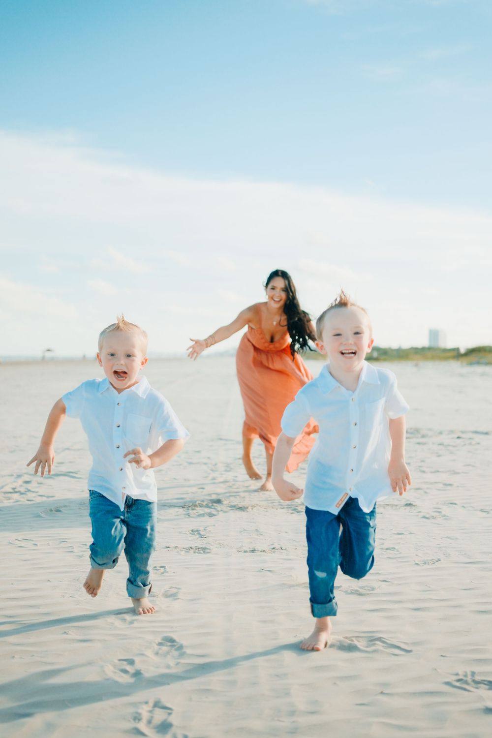 Two joyful boys in white shirts and jeans run on a sunny beach while a woman in an orange dress chases them.