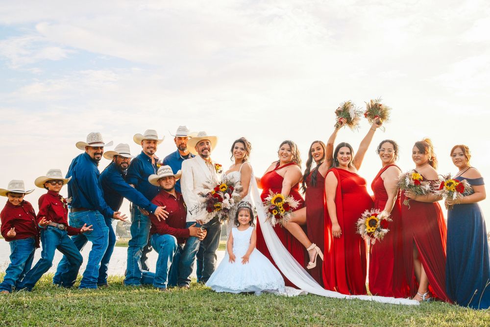 Wedding party in red dresses and blue suits with cowboy hats pose outdoors on green grass under a cloudy sky.