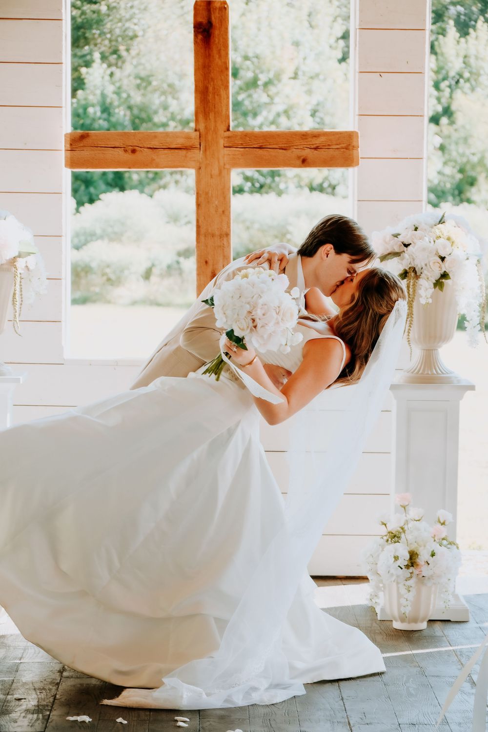 Bride and groom share a romantic dip kiss at the altar of a white chapel with a wooden cross.