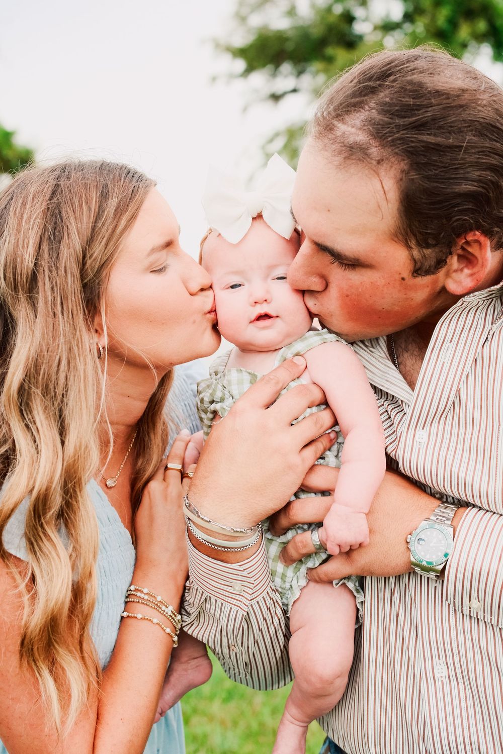 Two parents kissing their adorable baby girl on the cheeks outdoors in a sweet family portrait.