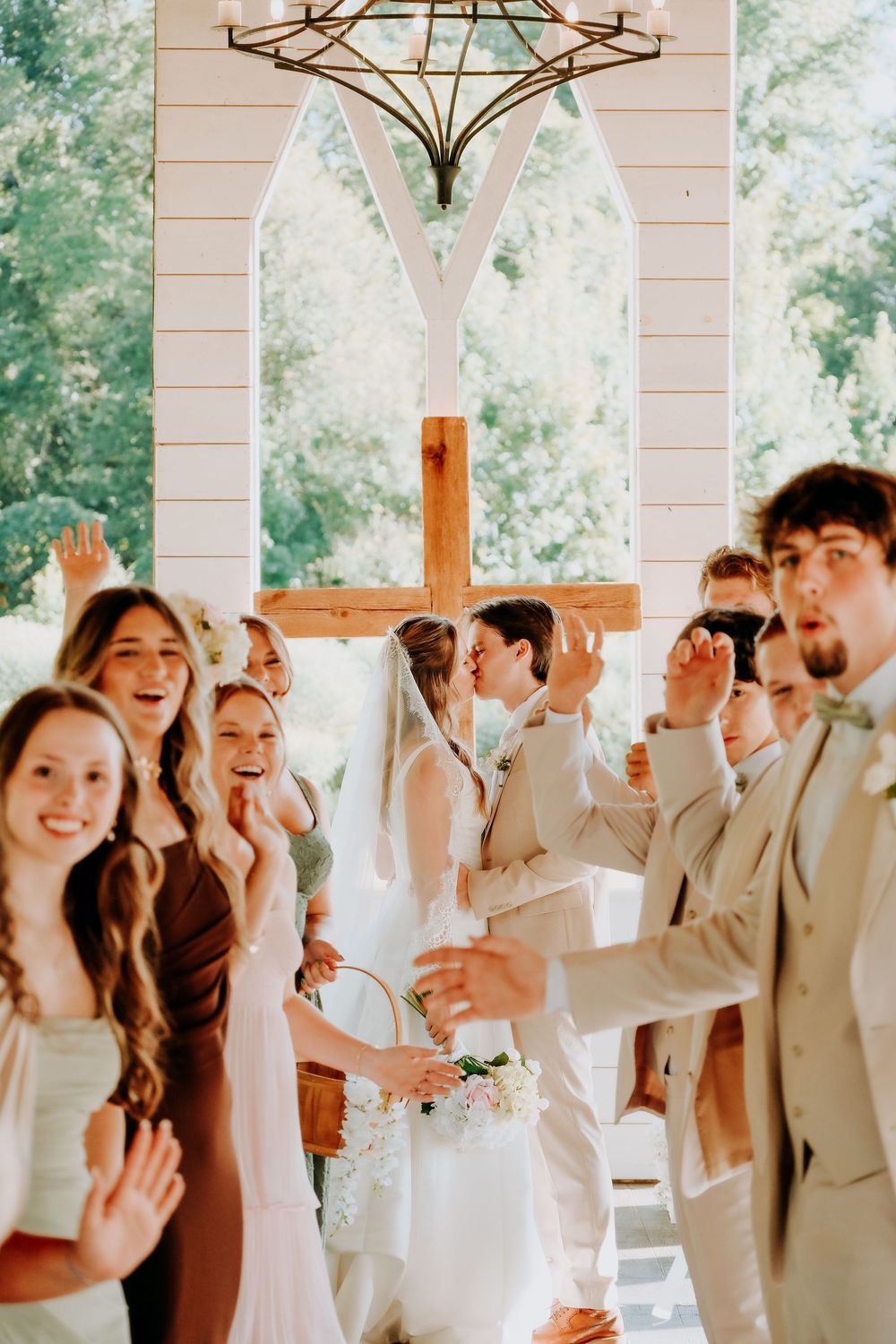 Wedding party cheers as bride and groom share first kiss beneath wooden cross in white chapel.