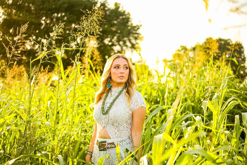 Blonde woman in white lace dress standing in tall golden sunlit grass.