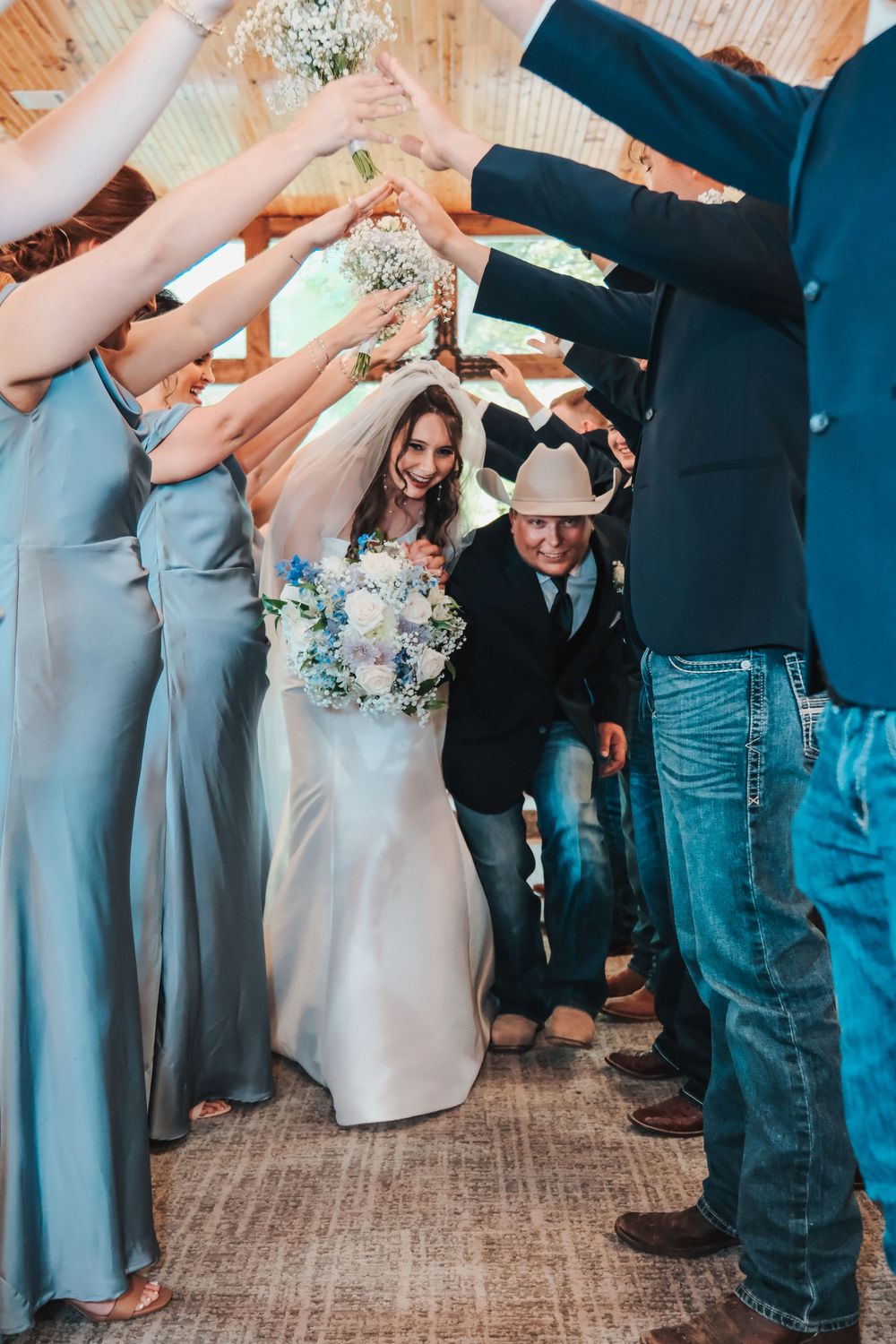 Bride and groom walk through arch of raised hands formed by wedding party in rustic barn venue.