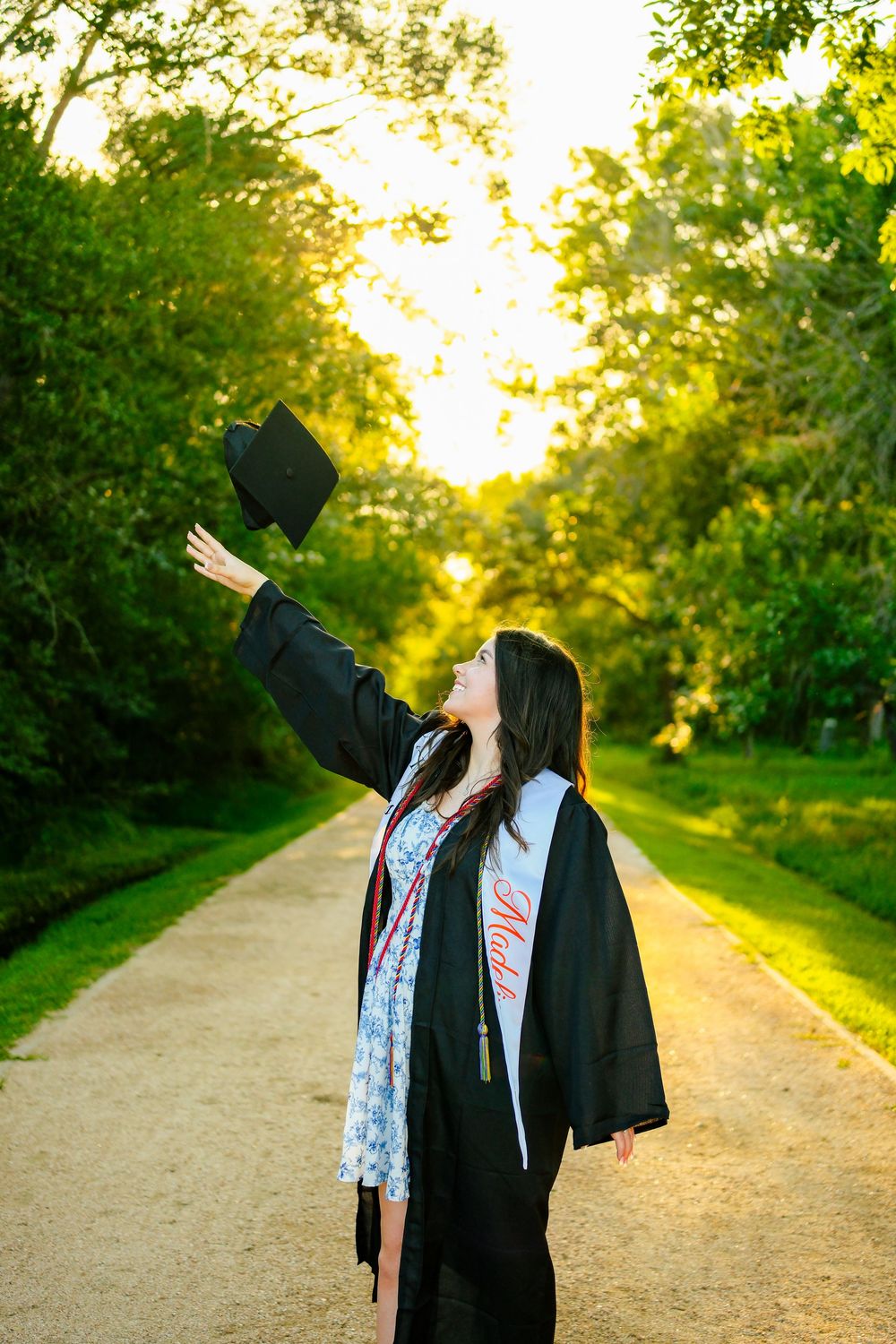 A young woman in graduation cap and gown tosses her mortarboard in the air on a sunlit tree-lined path.
