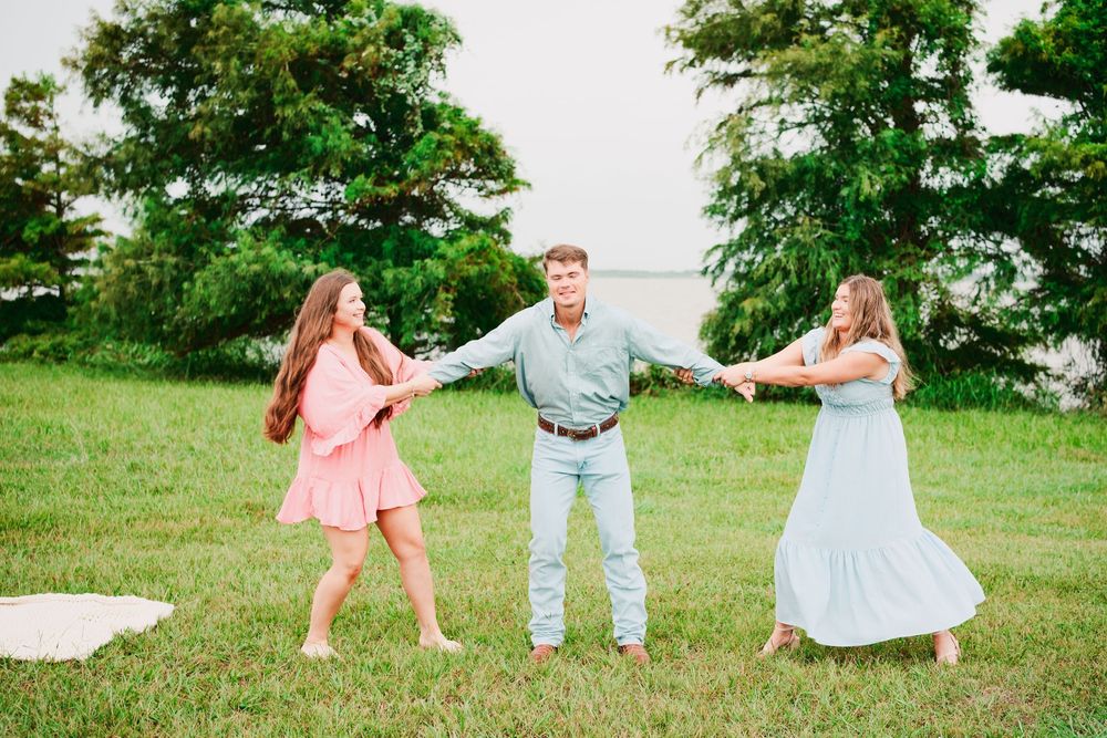 A man in a blue shirt and jeans holds hands with two women in pastel dresses while dancing outdoors on green grass.