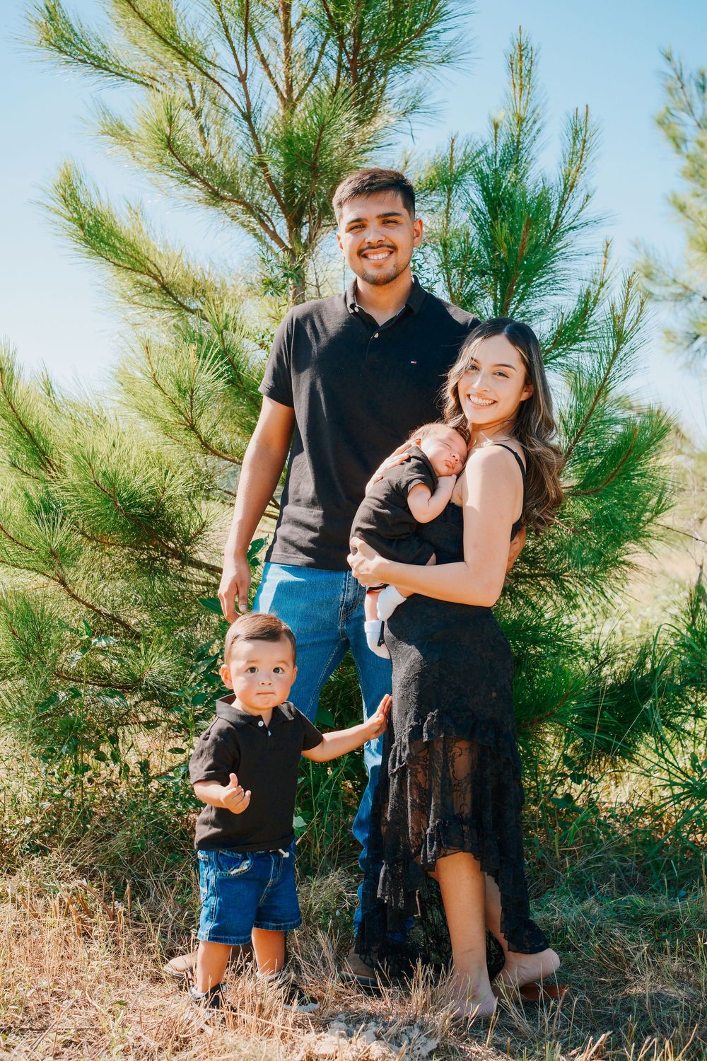 Young family of four poses outdoors near pine trees, parents in black outfits with toddler son and newborn baby.