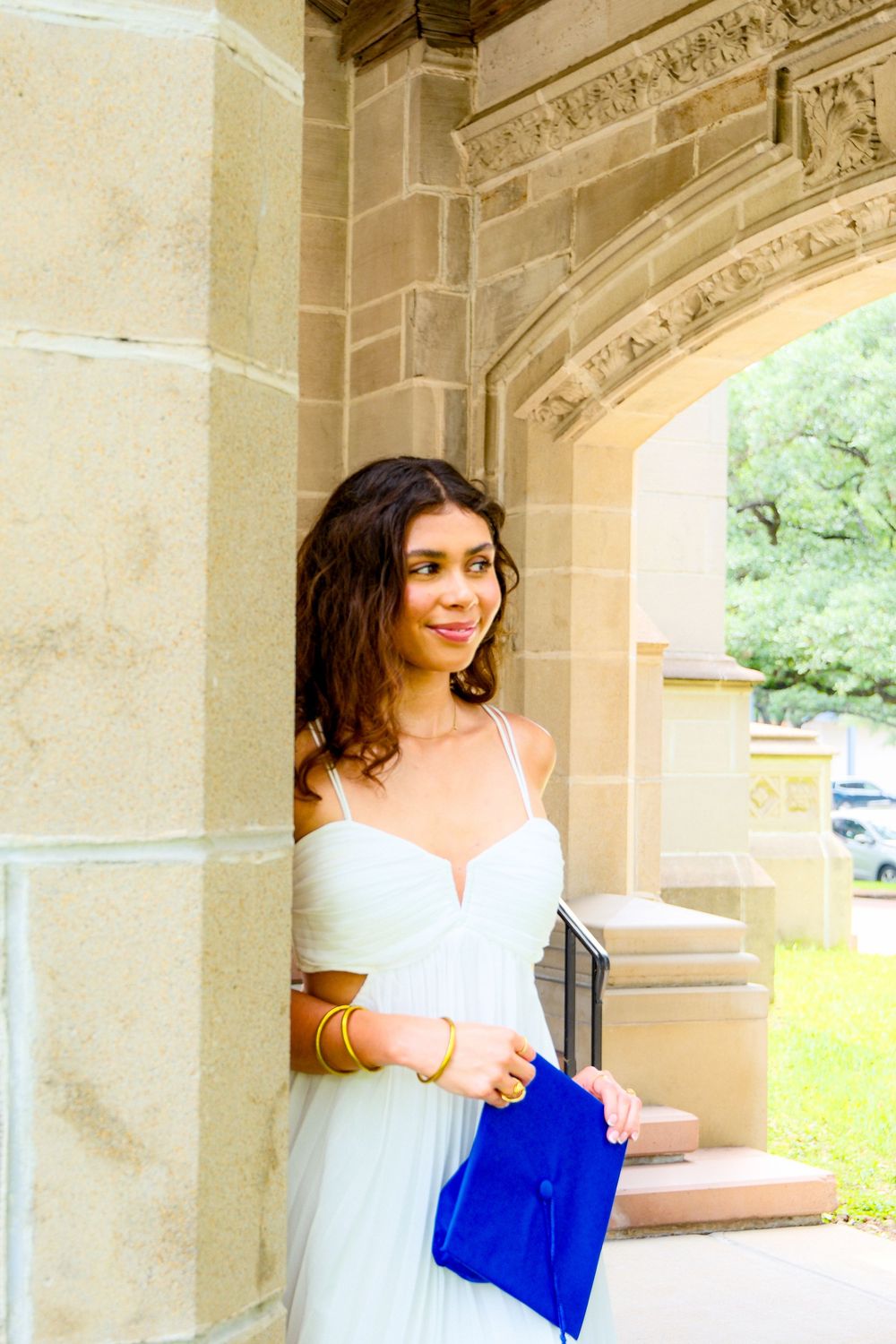 Young woman in white dress holding blue graduation cap by stone archway.
