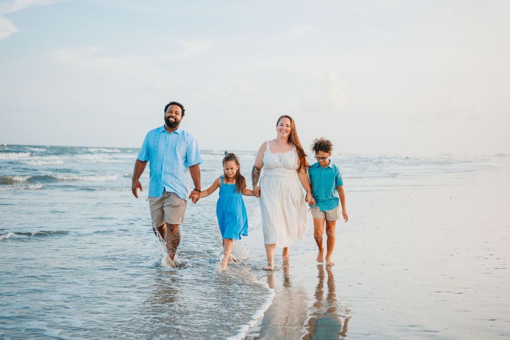 Family of four walking hand-in-hand along a sunny beach shoreline, parents with two young children in matching blue outfits.