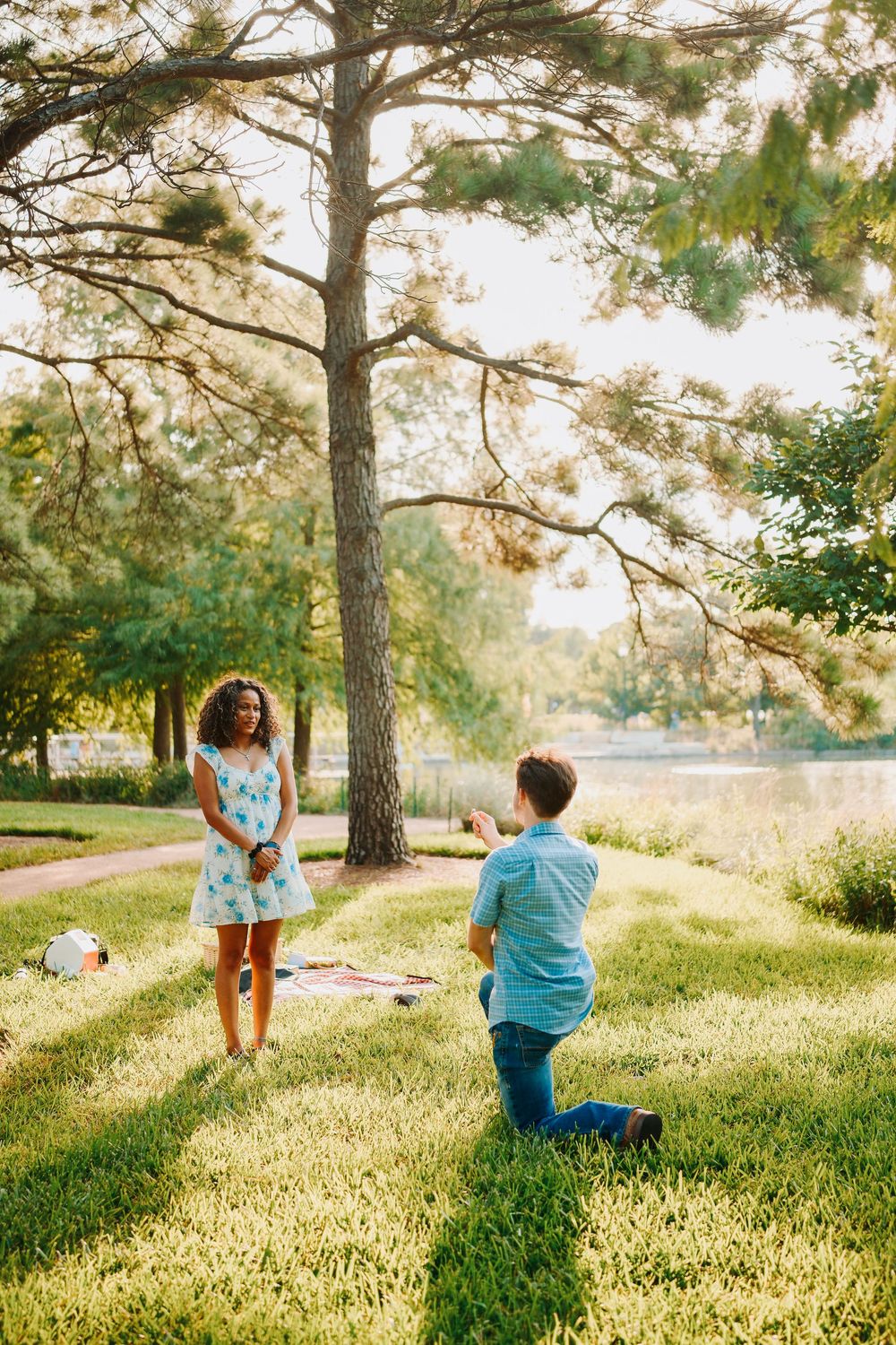 Man kneeling on one knee proposing to woman in blue dress in sunny park near lake.