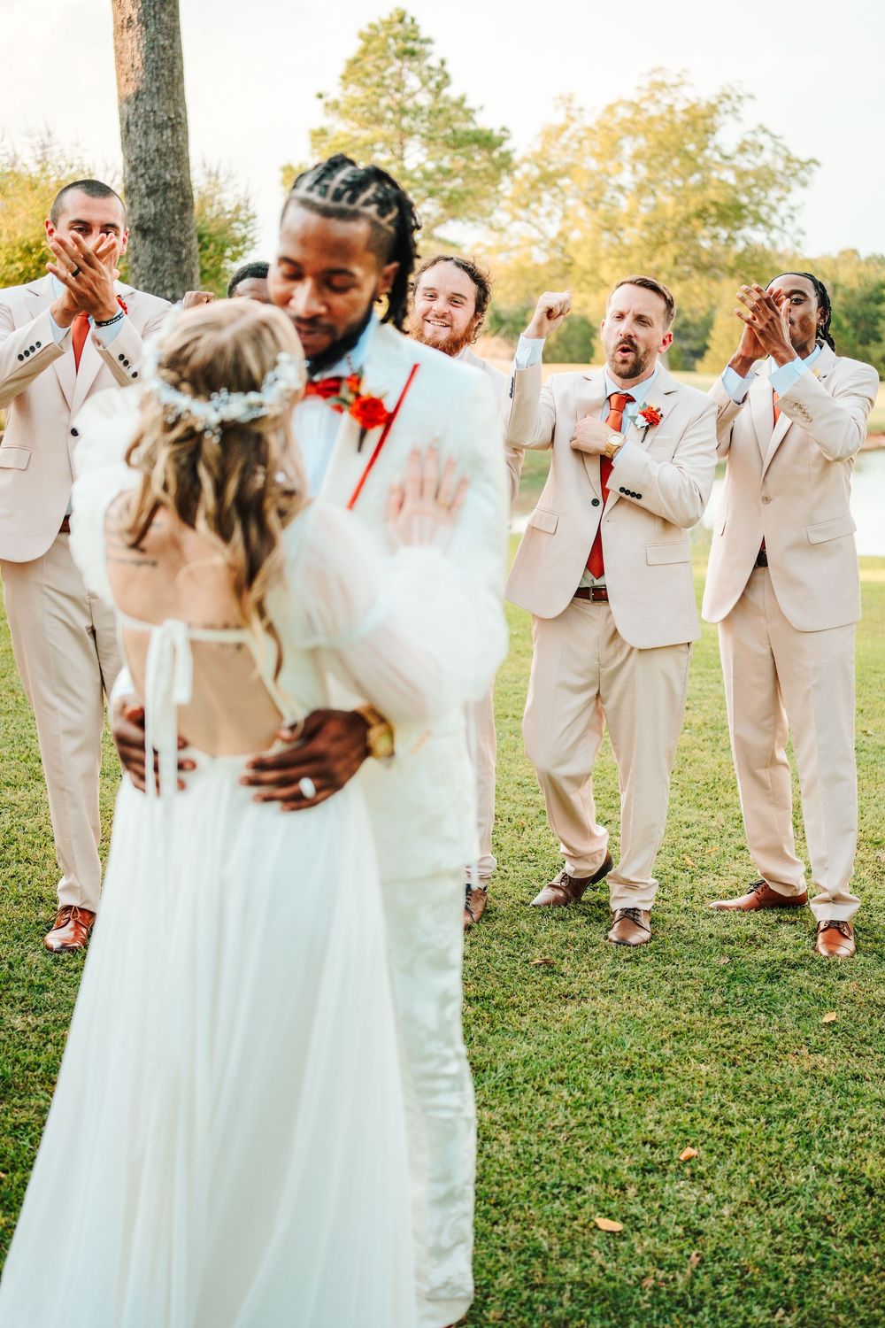 Groom embraces bride in white dress as groomsmen in tan suits celebrate joyfully outdoors at a wedding ceremony.