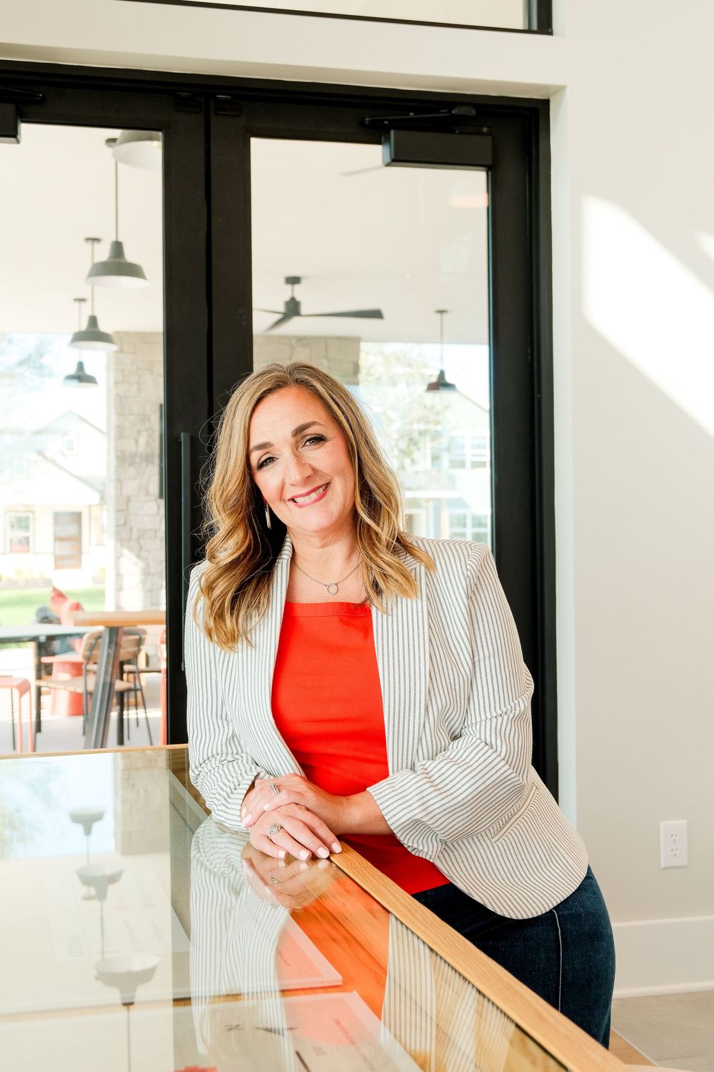 Smiling blonde woman in striped blazer leaning on glass railing indoors.