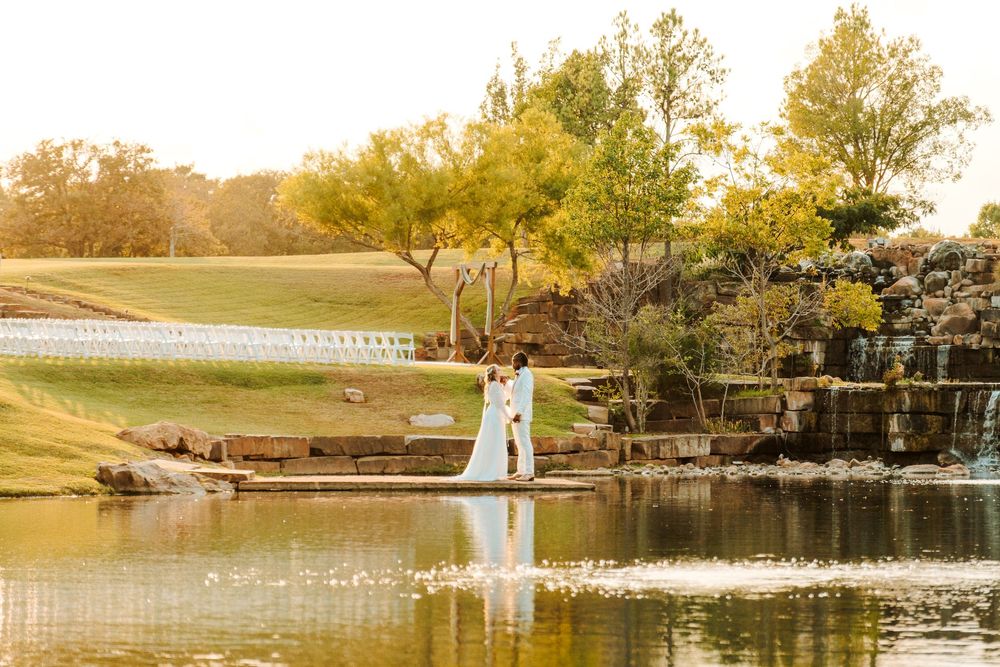 Bride and groom standing by a serene pond with a waterfall and lush trees in golden sunset light.