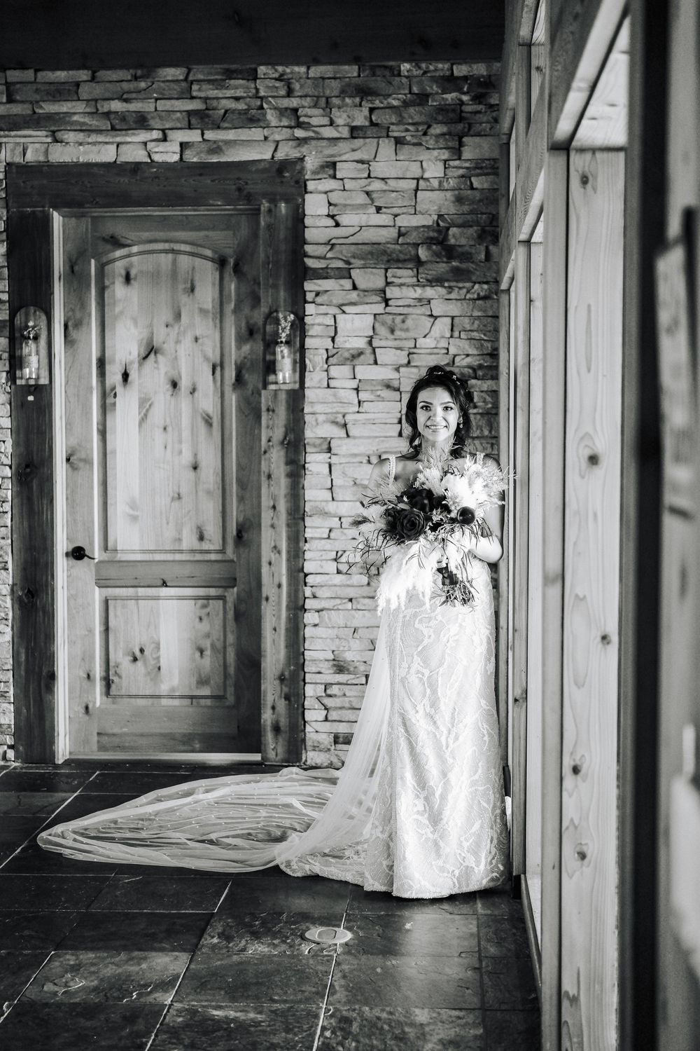Bride in lace wedding gown with long train holds bouquet beside rustic stone wall and wooden door.