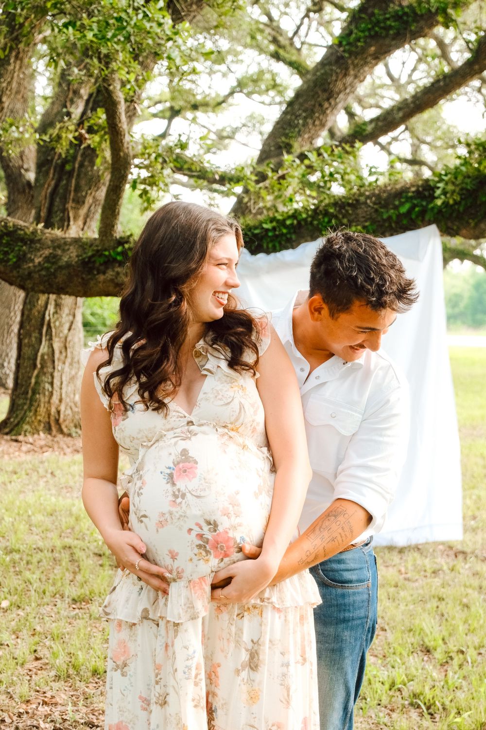Smiling pregnant couple standing under a large oak tree in a floral dress.