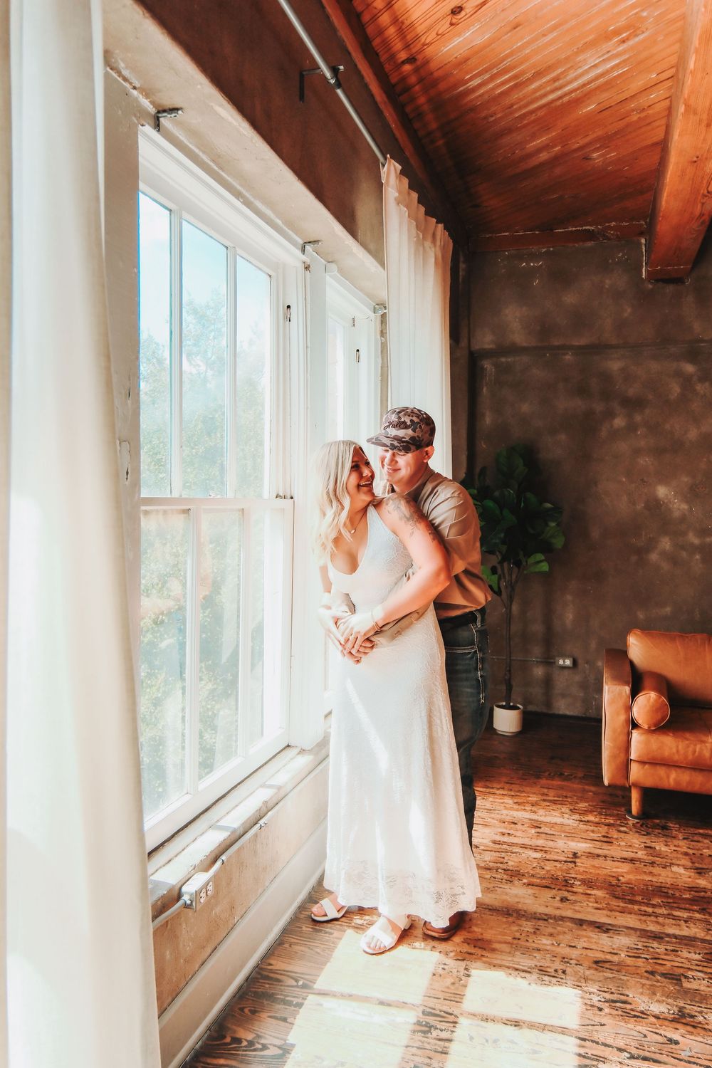 Couple embracing by sunlit window in rustic room with wood floors, ceiling, and leather chair, woman in white dress.