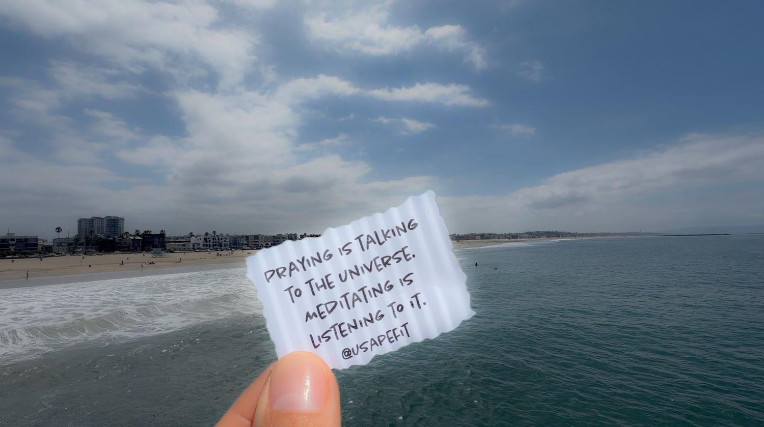 Hand holding a note about prayer and meditation on a sunny beach with ocean views and blue skies.