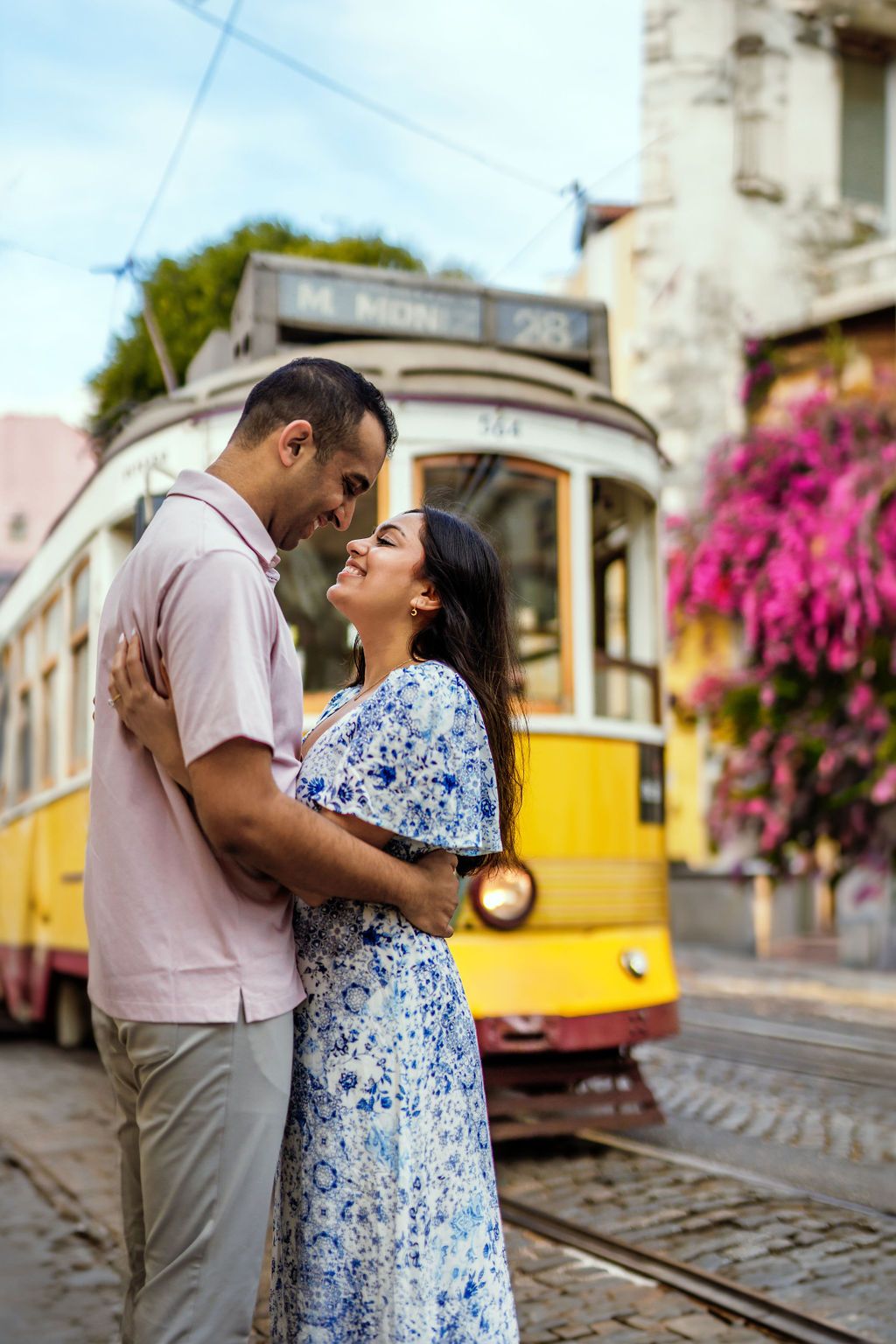 Couple holding hands on cobblestone street beside a yellow Lisbon tram, with a historic church tower in the background.