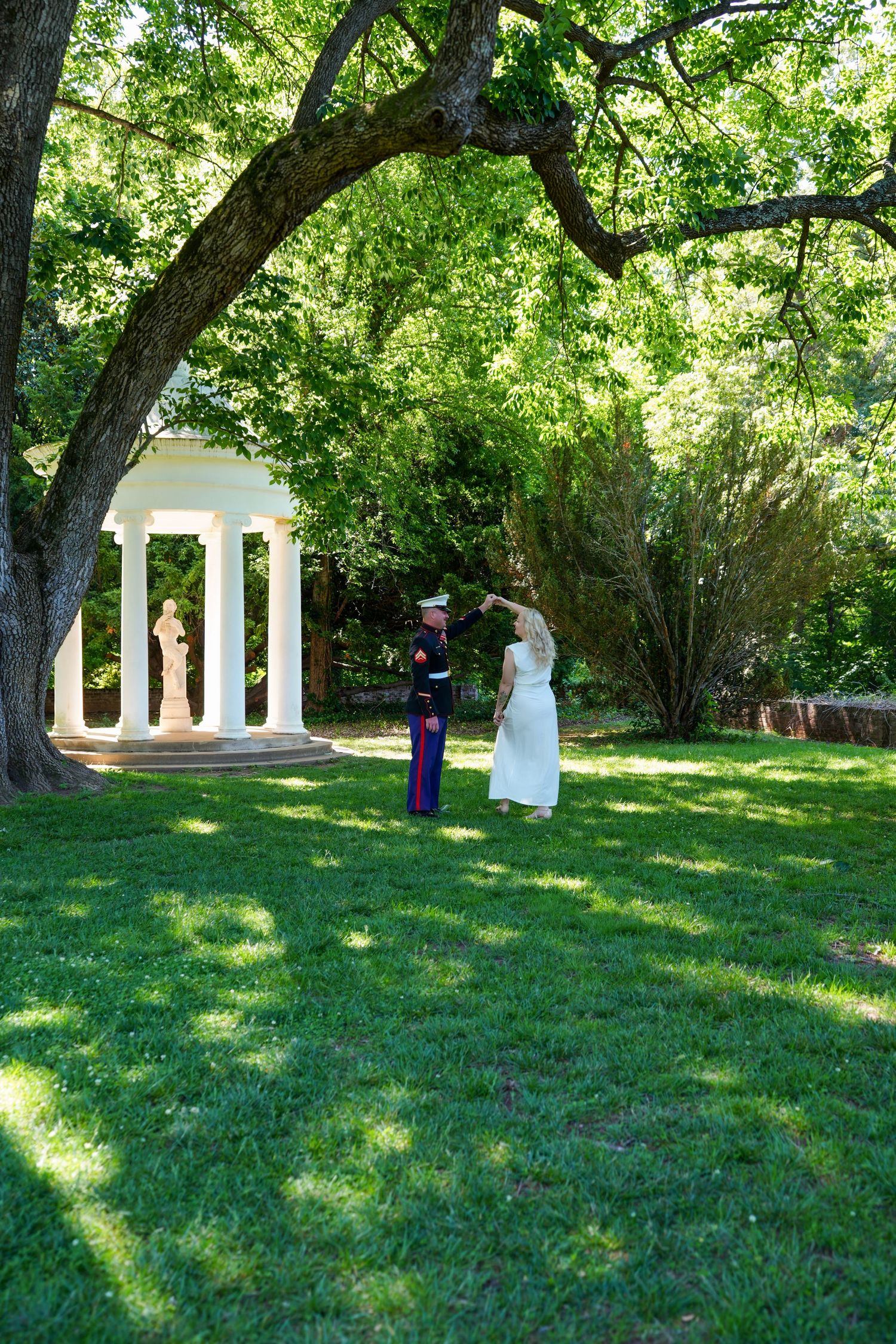 Couple embracing in a lush green garden park with a white columned gazebo and statue in the background.