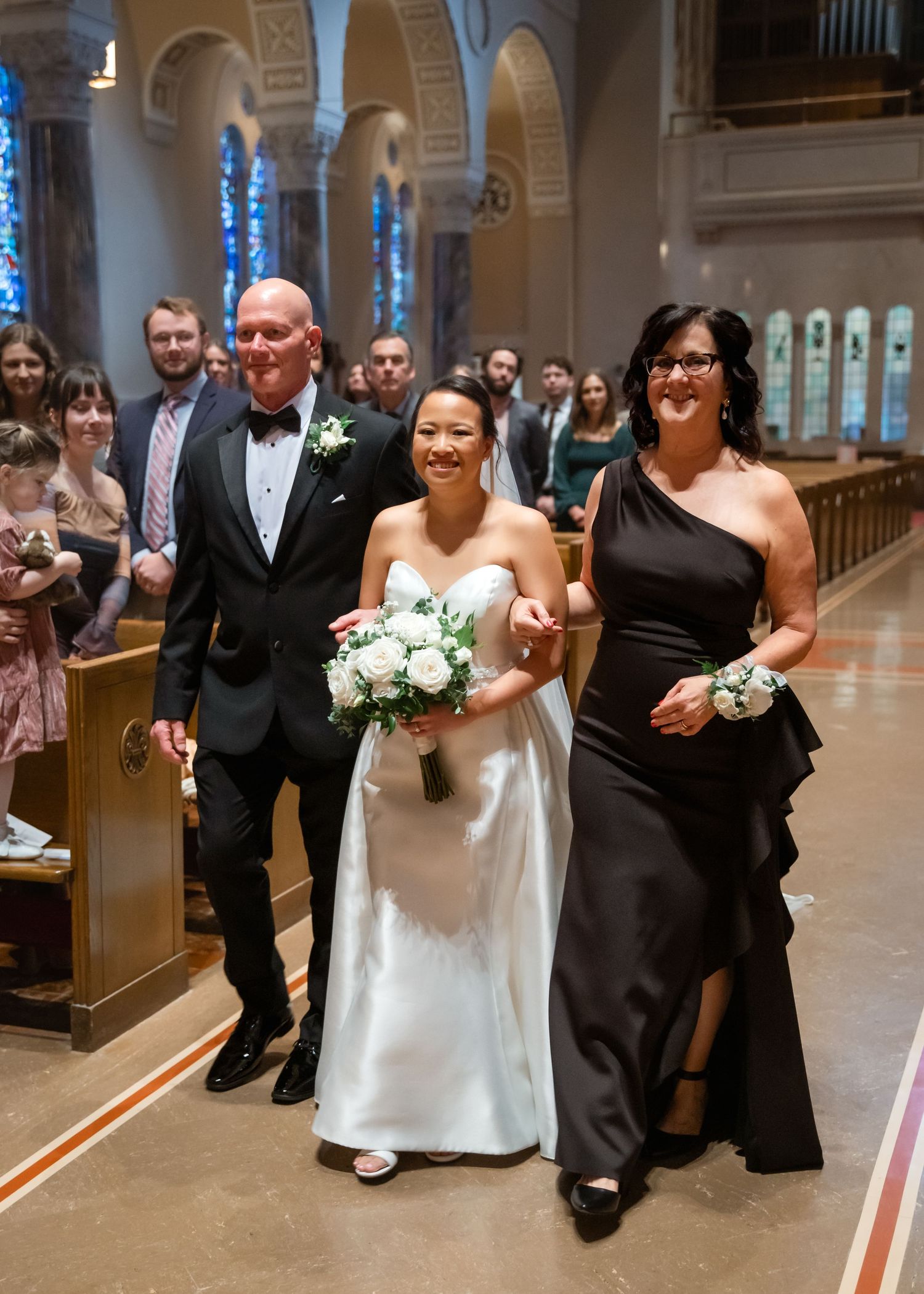 Bride walks down church aisle in white gown holding bouquet, escorted by man in tuxedo and woman in black dress.