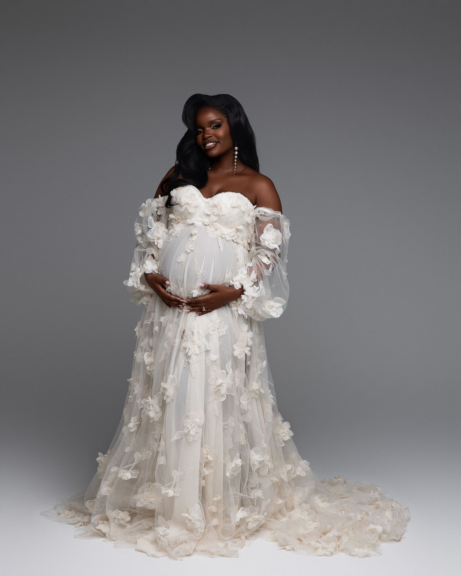 Pregnant woman in elegant off-shoulder white floral lace maternity gown smiling during a studio photoshoot.