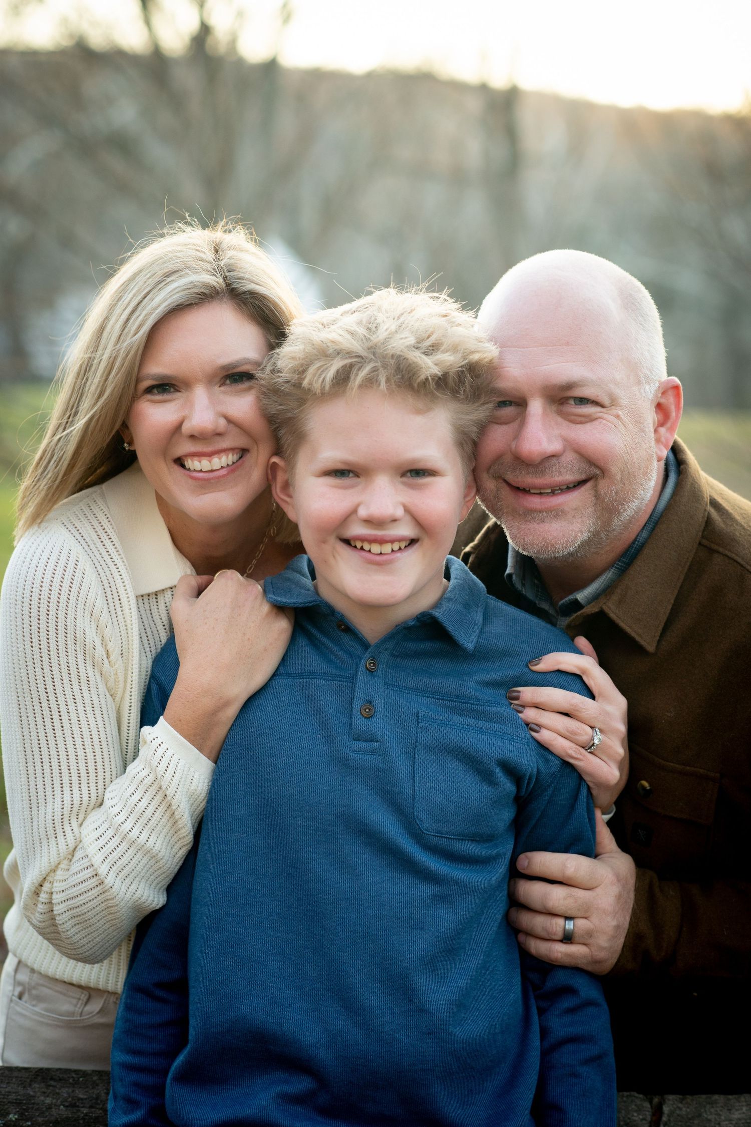 A smiling family of three poses outdoors, featuring a blonde woman, a young boy in blue, and a bald man in brown jacket.
